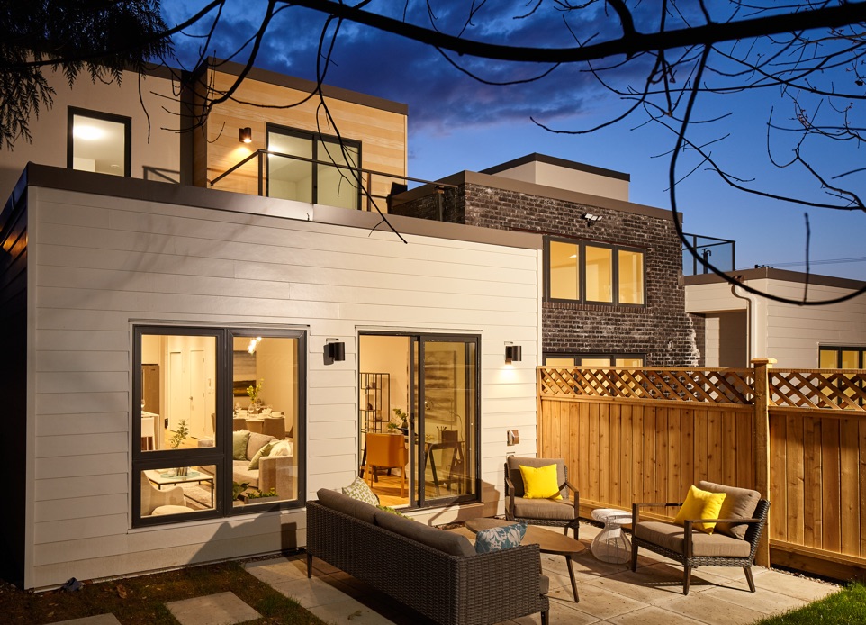 Exterior image looking to the living space from the back yard and second entrance from the back patio featuring white horizontal cladding, designed by N B A architects. Cozy exterior patio furniture with yellow pillows and a wooden fence behind.