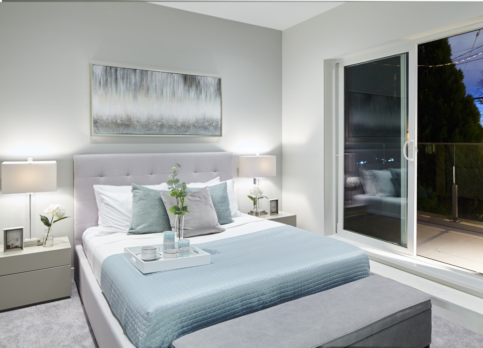 Interior image of a master bedroom featuring high ceilings and a cozy queen bed with white interior with light blue elements, featuring a wide balcony with sliding door.