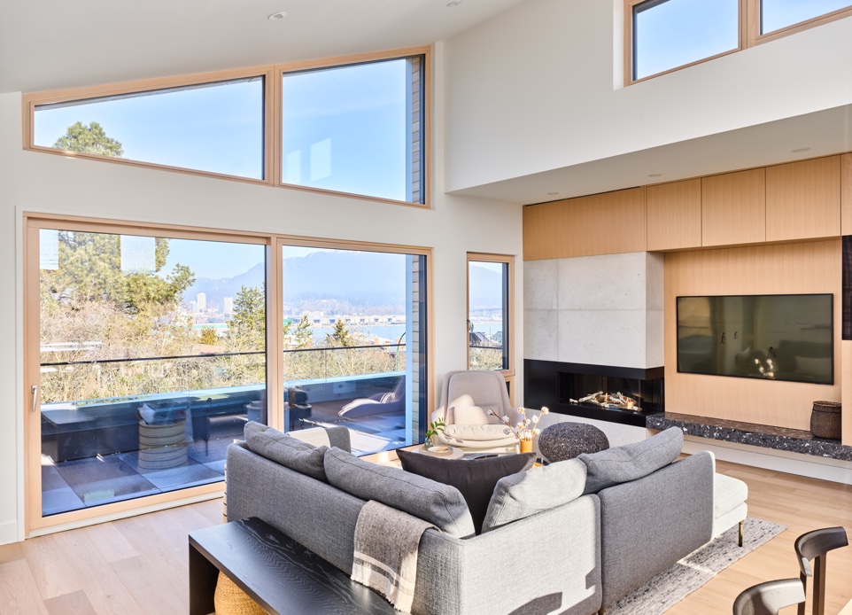 The interior image of the living room featuring the spacious outdoor patio space with a spectacular water panoramic view and the cozy fireplace with white and light wood elements. The upper floor contains a large open-plan living area with a 12’ high cathedral ceiling, seamlessly opening out onto a large upper view deck.