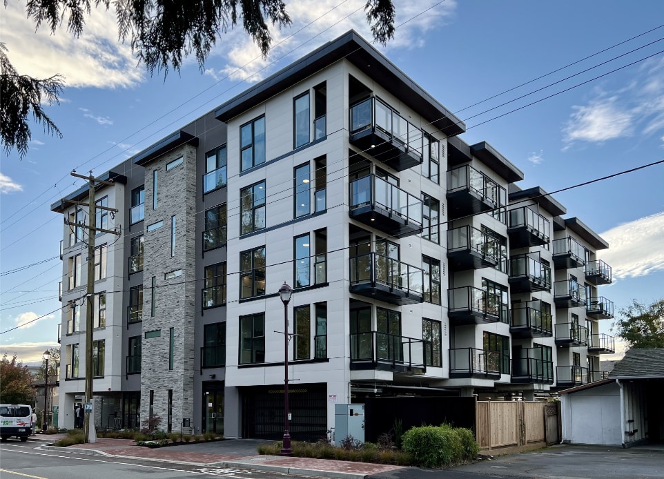 The exterior image of completed modern mid-rise, 6-stories strata apartment building above a podium car park ground parking in Langford, BC, designed by nba architects. Featuring rooftop resident amenity space opening onto a rooftop garden. 