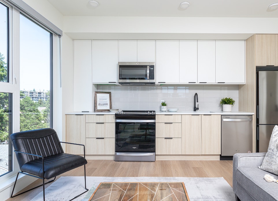 The interior image of the 1 bed apartment kitchen with plenty of natural light and selected interior furniture in a completed modern mid-rise, 6-stories strata apartment building above a podium car park ground parking in Langford, BC, designed by nba architects. Featuring rooftop resident amenity space opening onto a rooftop garden.