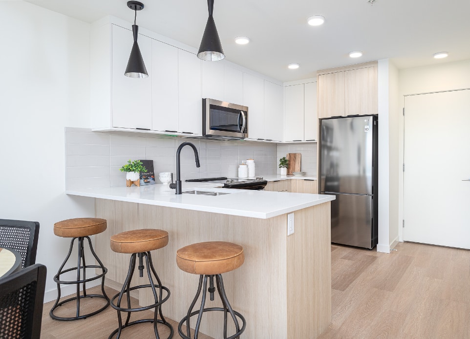 The interior image of the 1 bed apartment  kitchen with plenty of natural light and selected interior furniture in a completed modern mid-rise, 6-stories strata apartment building above a podium car park ground parking in Langford, BC, designed by nba architects. Featuring rooftop resident amenity space opening onto a rooftop garden.